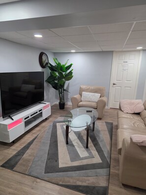 Ett vardagsrum med en platt-TV, ett soffbord i glas, en beige soffa, en krukväxt och en matta.