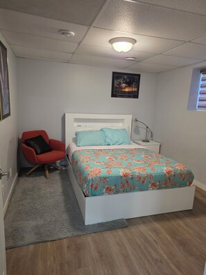 Ett sovrum med en säng, en röd stol, ett nattduksbord, en lampa och en inramad tavla på väggen.