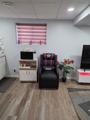 A modern living room with a black and red gym chair, a microwave, a small cabinet, a potted plant, and a television. - Spacious 2 bedroom basement in a quiet neighborhood. (Winnipeg)