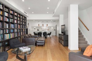 Ein modernes Wohnzimmer mit einer großen Bücherwand, Ledersofas, einem Glastisch und einem Essbereich mit Stühlen, im Hintergrund befindet sich eine Küche.