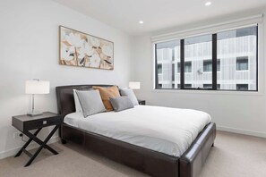 A modern bedroom with a large bed, two bedside tables, a wall-mounted lamp, and a large window with a view of a building.