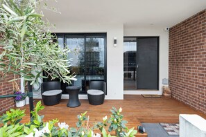 A modern outdoor patio with a wooden deck, two black wicker chairs, and a large potted plant.