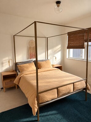 Een hemelbed met baldakijn, een nachtkastje met lamp, een aan de muur bevestigde dromenvanger, en een raam met jaloezieën.