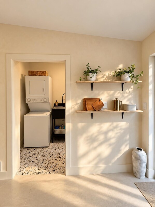 Een wasruimte met een wasmachine, droger en een klein tafeltje met een mand. Boven het tafeltje staan twee planken met planten en keukenspullen.