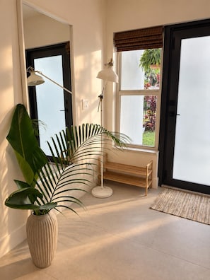 Une pièce dotée d’un grand miroir, d’une plante en pot, d’un lampadaire, d’un banc et offrant une vue sur un jardin par une fenêtre.