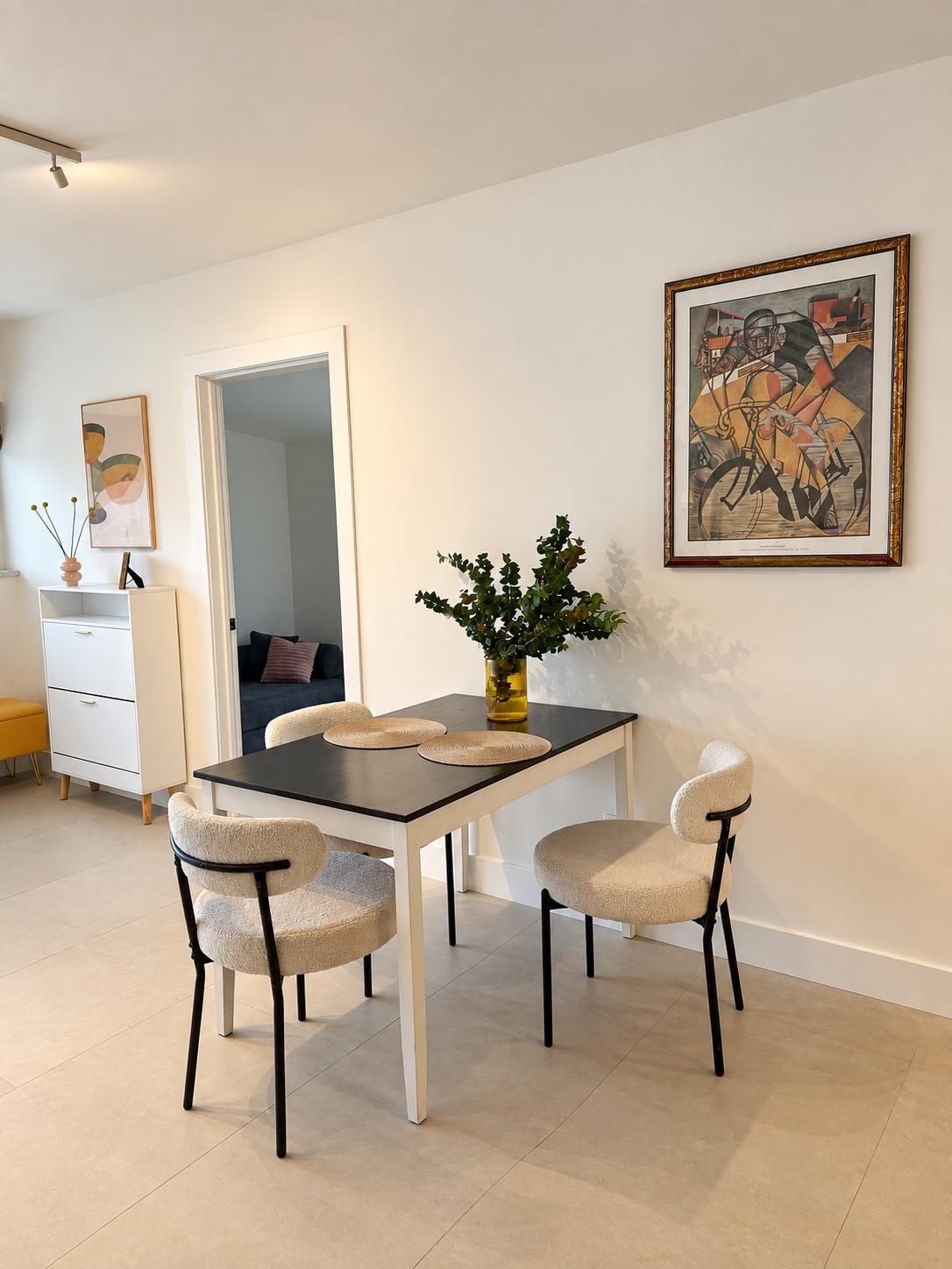 Ein Essbereich mit Tisch, Stühlen und einer Vase mit Grünpflanzen. An der Wand hängt ein gerahmtes Bild und eine Tür führt in einen anderen Raum.