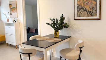 Ein Essbereich mit Tisch, Stühlen und einer Vase mit Grünpflanzen. An der Wand hängt ein gerahmtes Bild und eine Tür führt in einen anderen Raum.