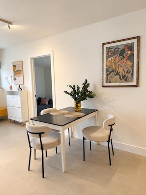 Un coin repas avec une table, des chaises et un vase agrémenté de plantes vertes. Un tableau encadré orne le mur, et une porte donne accès à une autre pièce.