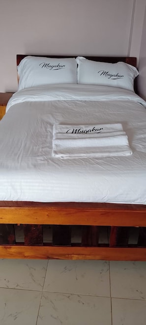 A neatly made bed with a white bedspread and pillows, a folded white towel with the word 'Mugakar' on it, and a wooden bed frame. - Hotel Delite (Chuka)