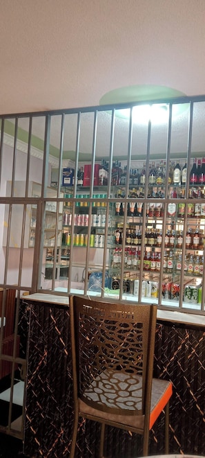 A bar area with shelves stocked with various bottles and cans, a metal partition, and a chair with a patterned backrest.