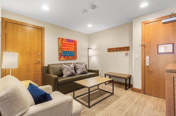 Ein möbliertes Wohnzimmer mit einer Couch, einem Couchtisch und einer Bank. Eine Holztür befindet sich rechts, und eine Wandlampe ist angebracht.