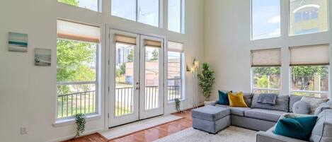 A spacious living room with a grey sectional sofa, a white rug, and large windows offering a view of the outdoors.