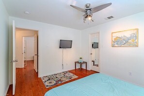 A bedroom with a bed, a ceiling fan, a flat-screen TV, a painting, and a door leading to another room. - Sleeps 16 | 2 Adjacent EaDo Homes Near Stadium (Houston)