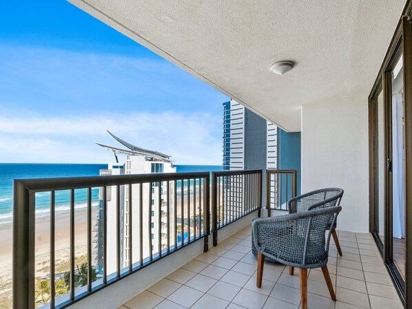Varanda com vista para a praia e o oceano, duas cadeiras de vime e um edifício moderno ao fundo.