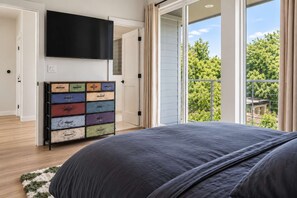 A bedroom with a large bed, a flat-screen TV mounted on the wall, a colorful dresser, and a balcony with a view of trees. - Regent Sol - Scandinavian Luxury - Central Austin (Austin)
