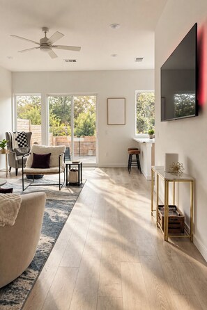 A spacious living room with a large window, a ceiling fan, a flat-screen TV, and a wooden floor. - Regent Sol - Scandinavian Luxury - Central Austin (Austin)