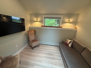Ein gemütliches Wohnzimmer mit einem an der Wand befestigten Flachbildfernseher, einem bequemen Sessel, einer Couch und einem kleinen Fenster mit Blick auf Grünflächen.
