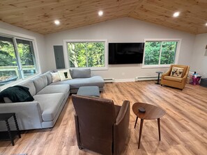 A modern living room with a sectional sofa, a brown leather chair, a round table, and a flat-screen TV mounted on the wall. - Tree top views, 1 mile to Mt Snow, spacious getaway , family & pet friendly (Dover)