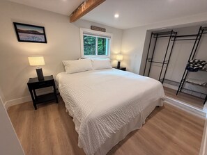 Ein Schlafzimmer mit einem Bett, zwei Nachttischlampen, einem Fenster, einem Kleiderhaken und einem gerahmten Bild an der Wand.