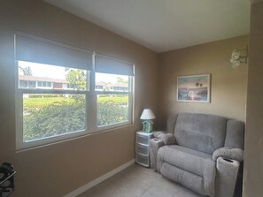 A room with a large window, a grey sofa, a small table with a lamp, and a painting on the wall. - Relax & Unwind in Englewood at Quails Run, over 55 community in Englewood. (Englewood)