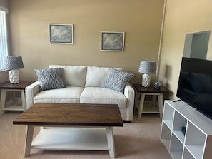A living room with a sofa, two side tables with lamps, a coffee table, a TV on a stand, and two framed pictures on the wall. - Relax & Unwind in Englewood at Quails Run, over 55 community in Englewood. (Englewood)