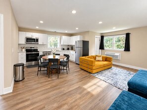 A modern kitchen with white cabinets, a dining table with chairs, a yellow sofa, and a blue couch. - Furnished Studio | 2 Full Beds | Short & Mid-Stay (Crystal Lake)