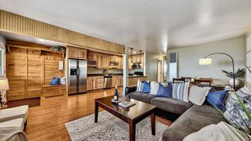 Uma cozinha moderna com armários de madeira e eletrodomésticos de aço inoxidável, uma sala de estar com sofá e mesa de centro, e uma sala de jantar com mesa e cadeiras.