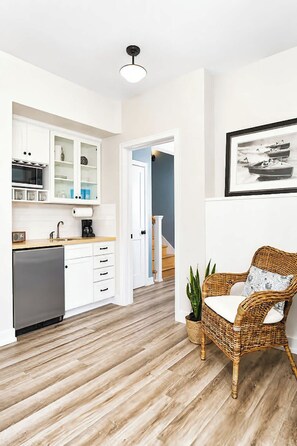 Eine moderne Küche mit weißen Schränken, einer eingebauten Mikrowelle und einer Kaffeemaschine. Ein Korbstuhl mit Kissen und ein Blumentopf sind ebenfalls vorhanden.