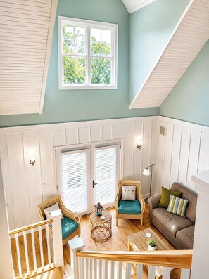 A spacious loft with a high ceiling, a large window with blinds, a cozy seating area with a brown sofa and wicker chairs, and a wooden staircase. - The Hampton | Pool • Hot Tub • Lake • Free Golf (Holland)