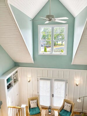 A room with a sloped ceiling, a ceiling fan, a large window with a view of the outside, two wicker chairs with cushions, and a small table with a lantern. - The Hampton | Pool • Hot Tub • Lake • Free Golf (Holland)
