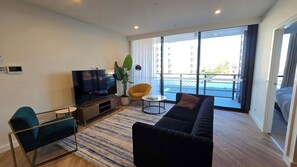 Ein modernes Wohnzimmer mit einem Flachbildfernseher, einer Couch, einem gelben Sessel und einem großen Fenster mit Blick auf einen Pool und Gebäude.