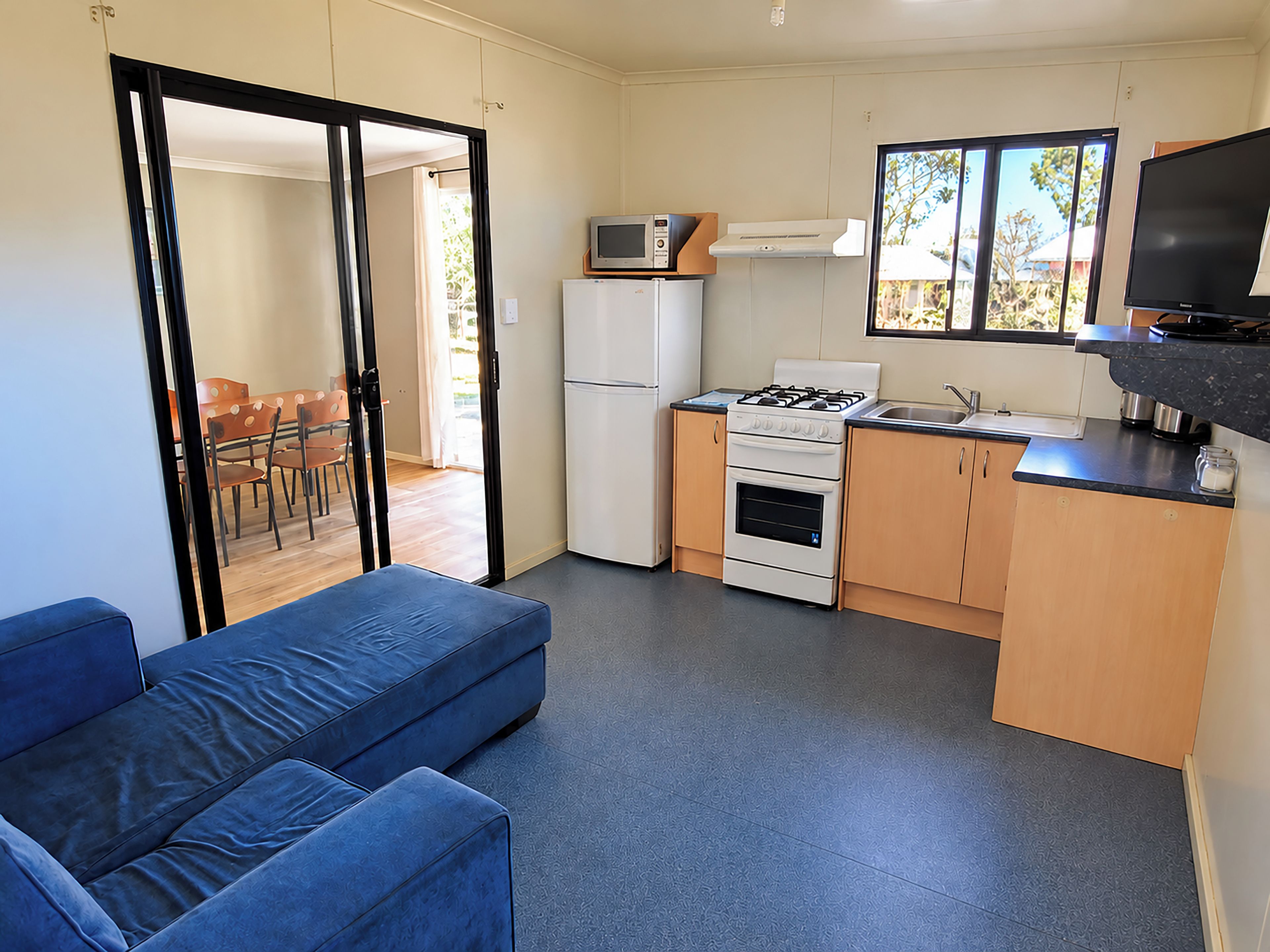 A compact kitchen with a gas stove, microwave, and refrigerator. There's a blue sofa and a dining area visible through an open door.