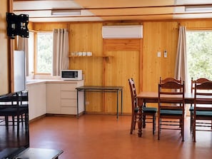 A compact room with a kitchenette, a dining table with chairs, a TV mounted on the wall, and a window with curtains.