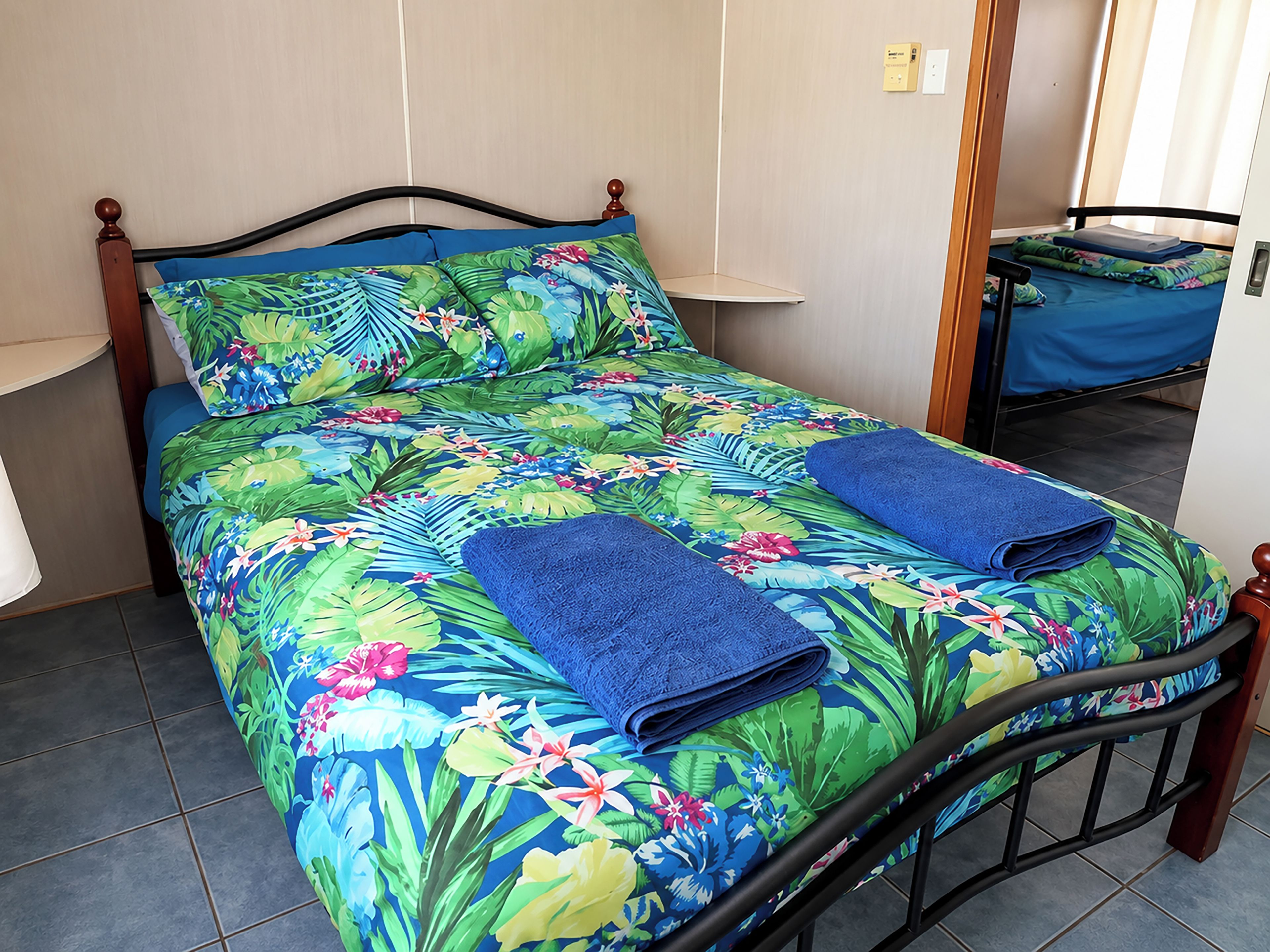 A bed with a floral bedspread and two folded blue towels, a wooden headboard, and a small table with a blue tablecloth.