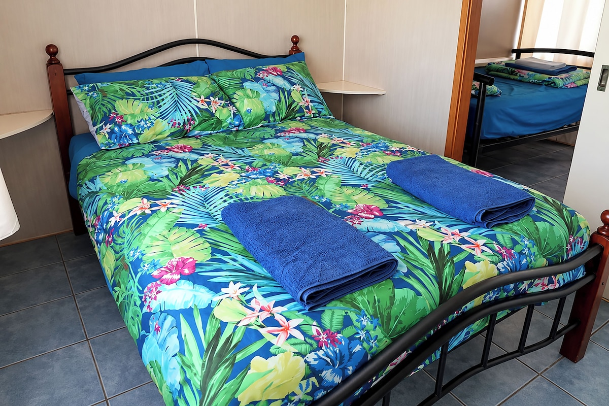 A bed with a floral bedspread and two folded blue towels, a wooden headboard, and a small table with a blue tablecloth.