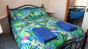 A bed with a floral bedspread and two folded blue towels, a wooden headboard, and a small table with a blue tablecloth.