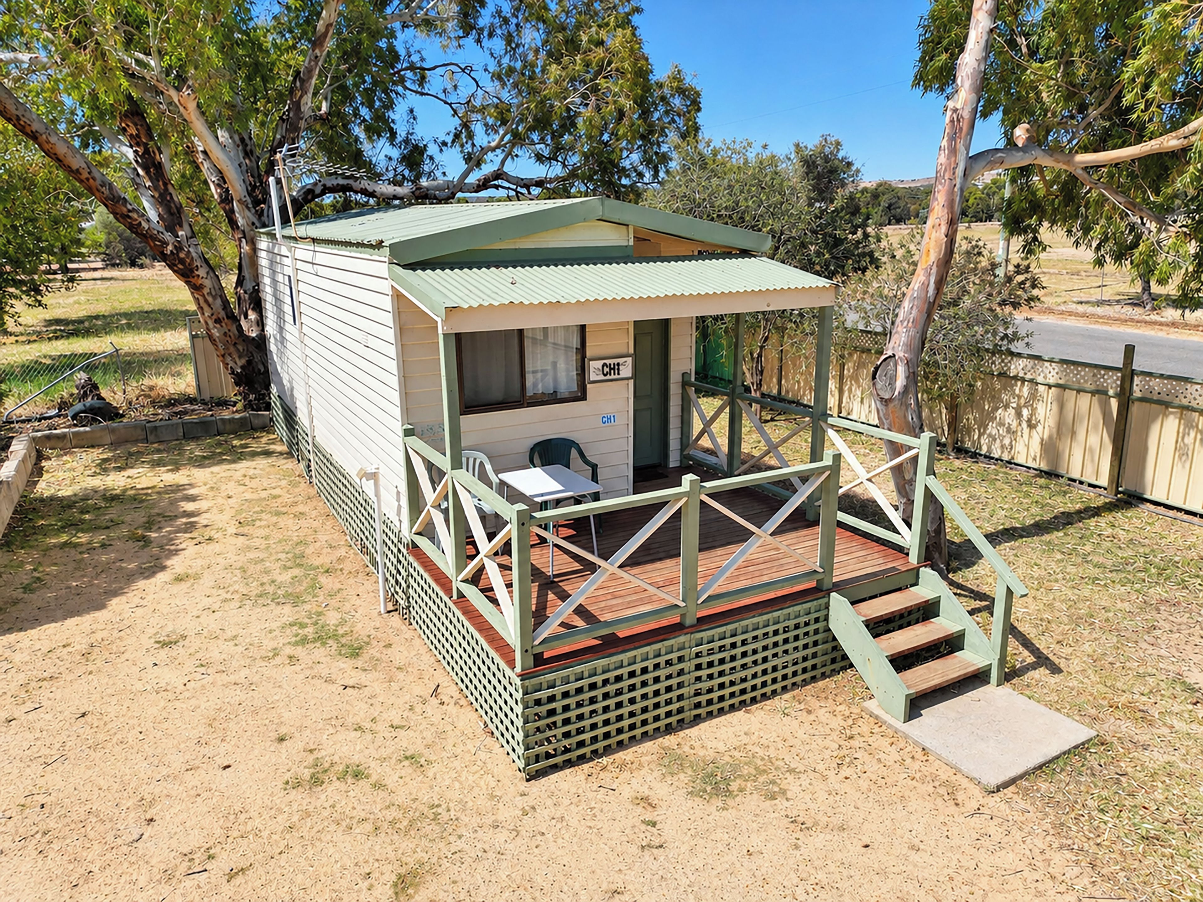 A small, single-story structure with a porch and a table, surrounded by trees and a fence.
