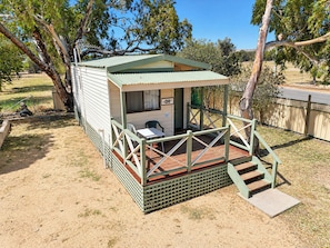 A small, single-story structure with a porch and a table, surrounded by trees and a fence.