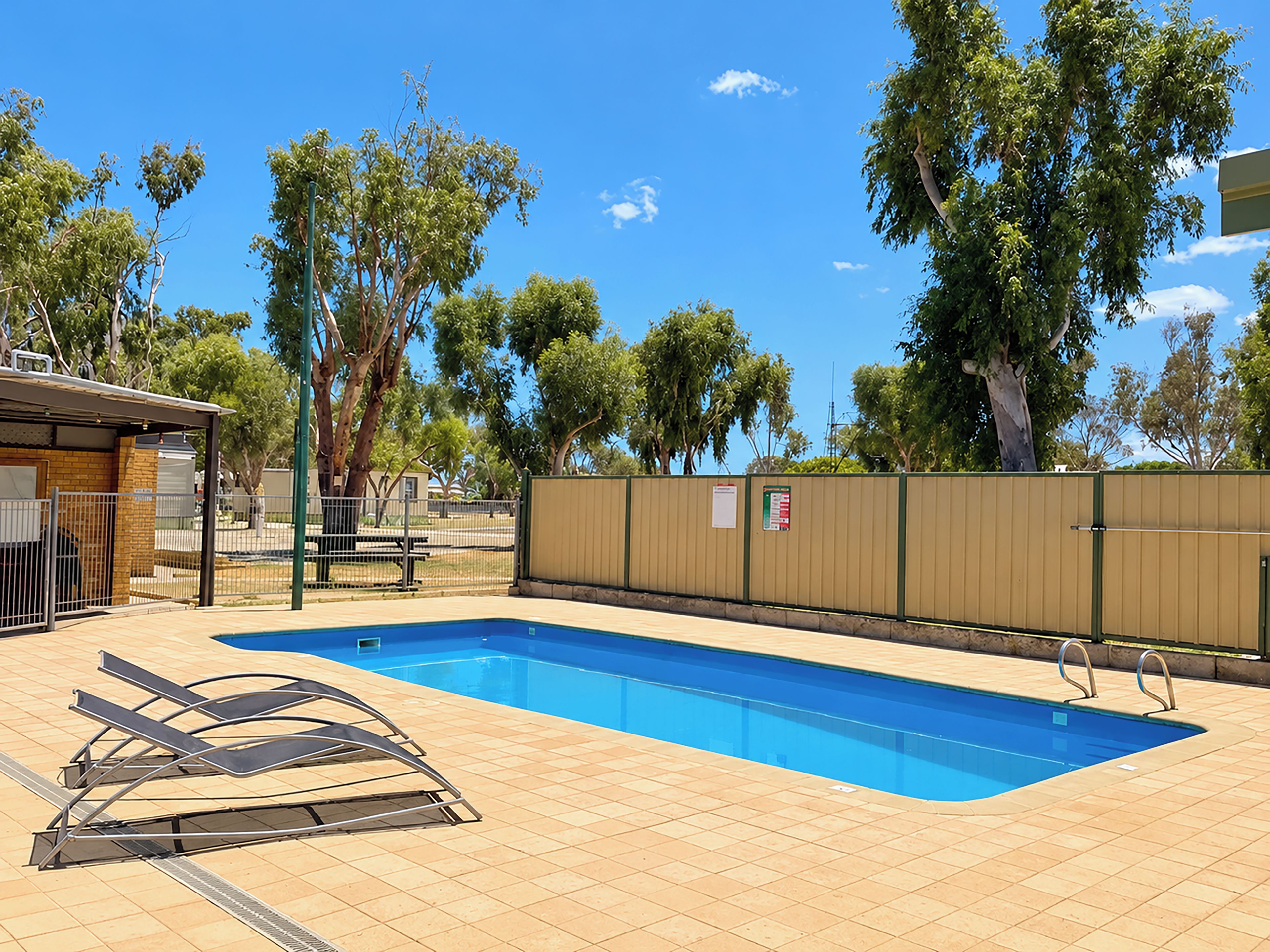 A swimming pool with a tiled deck, a fenced area, and trees in the background.
