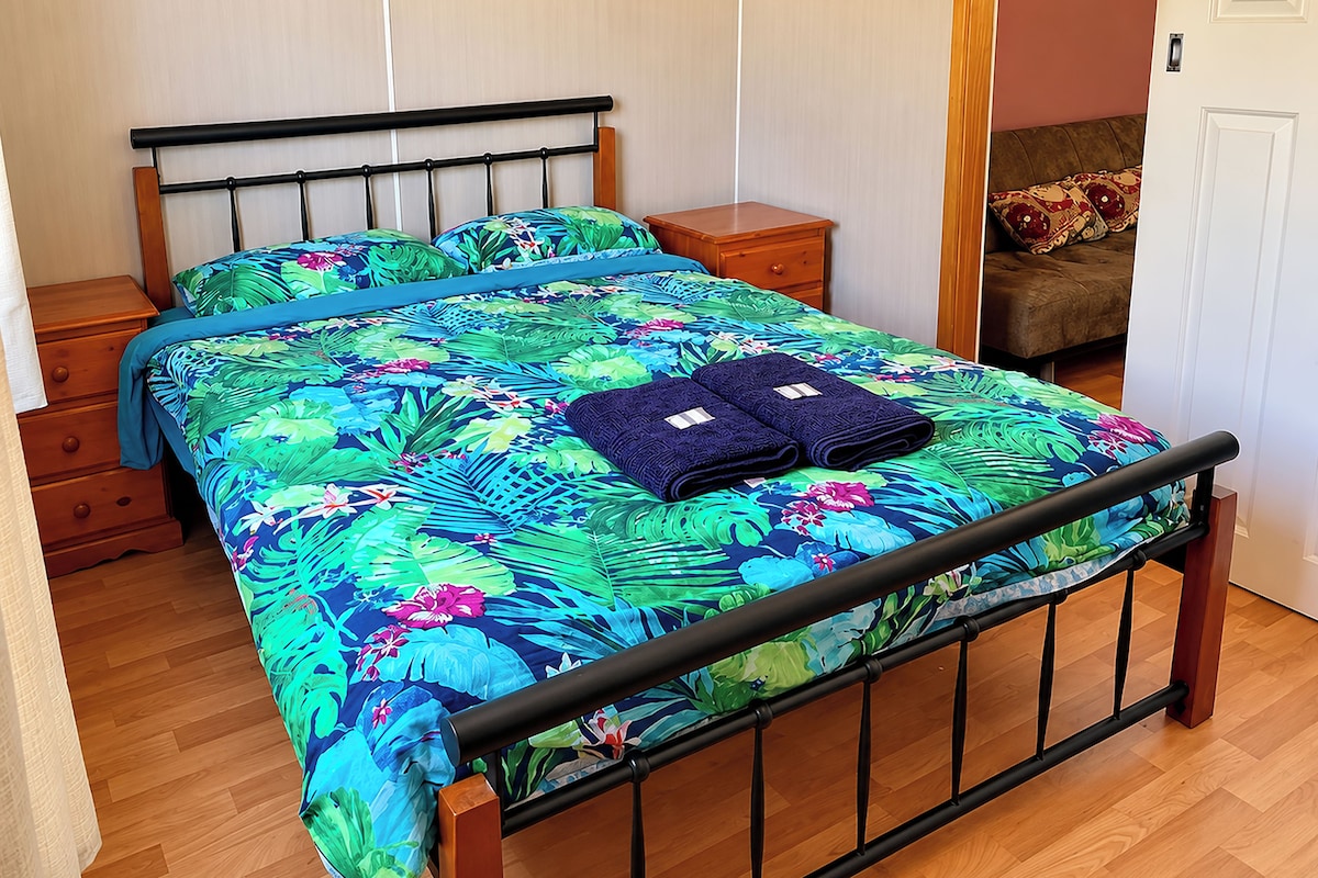 A bed with a floral bedspread, two folded towels, a wooden nightstand, and a wooden dresser.