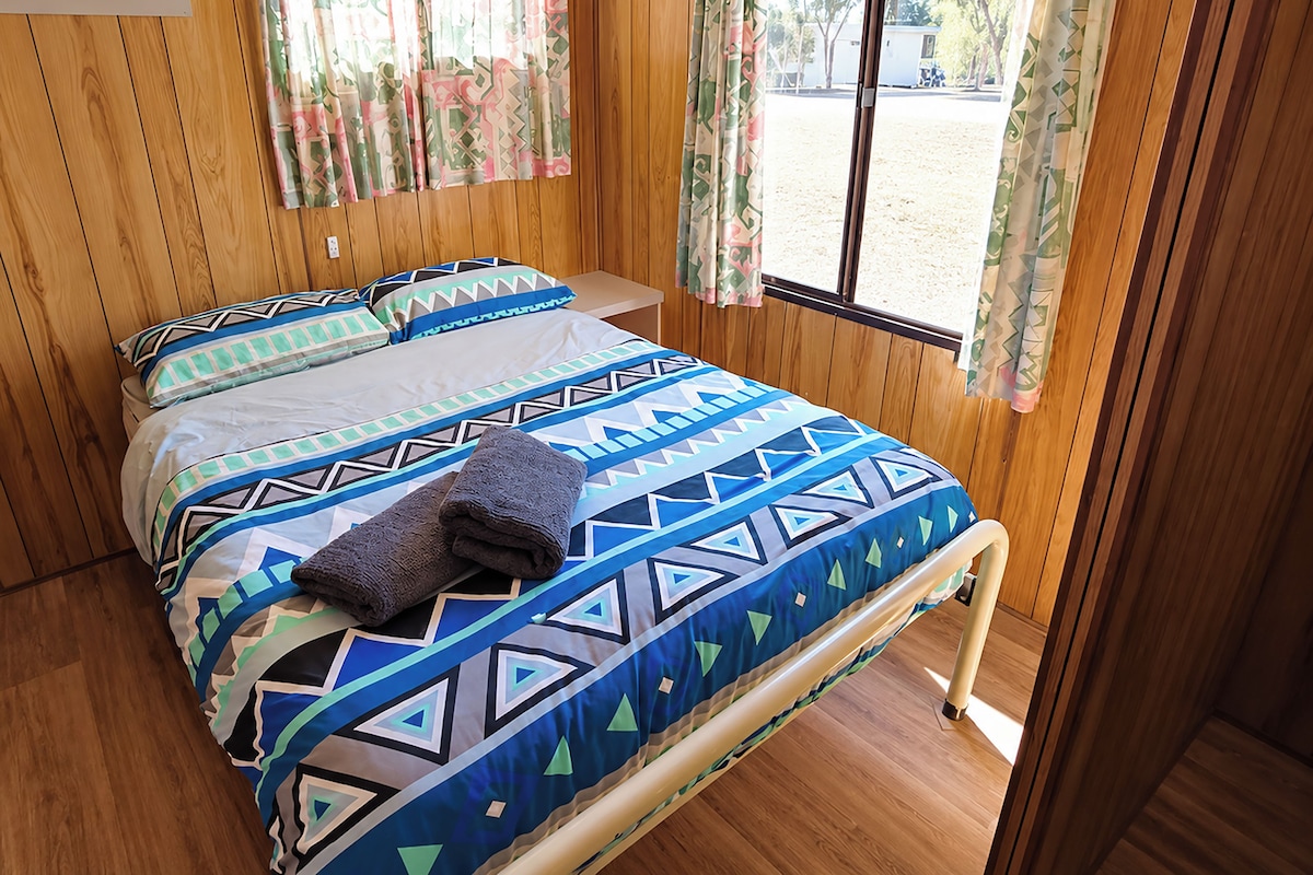 A room with a bed, wooden walls, a window with curtains, and a view of the outside.
