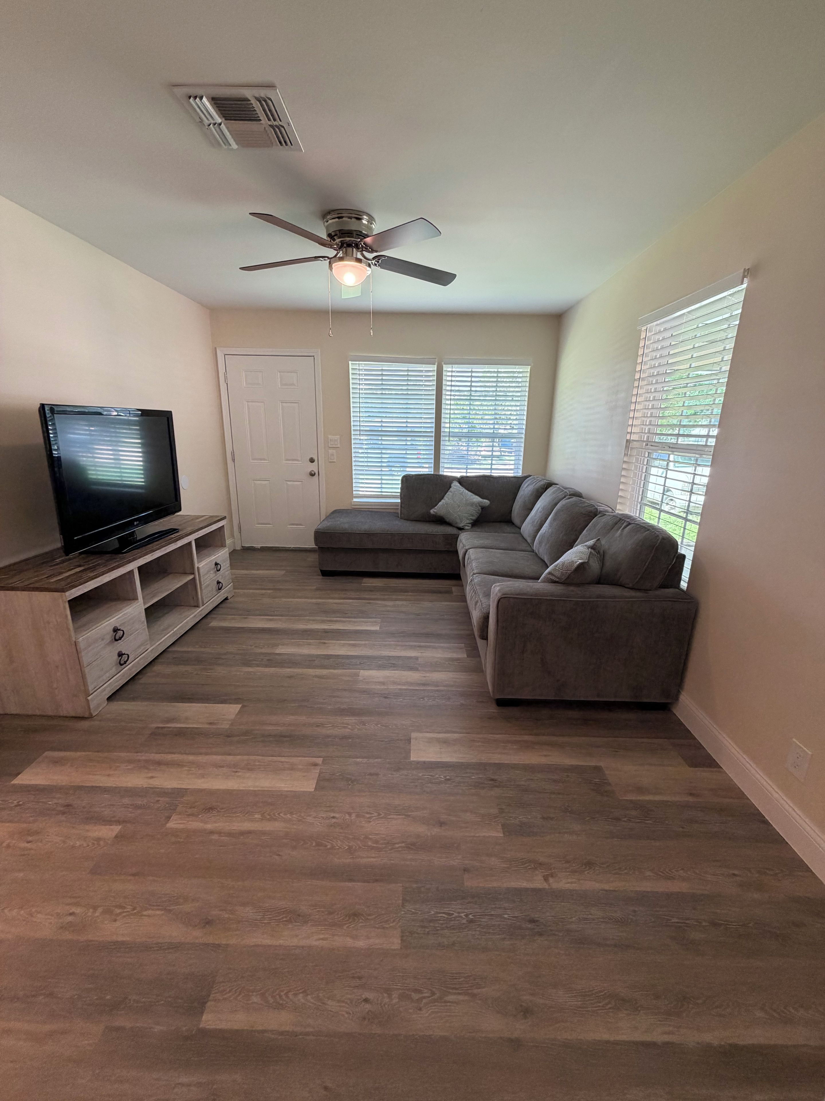 A living room with a flat-screen TV, a sofa, a ceiling fan, and a window with blinds.
