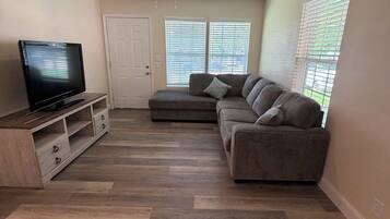 A living room with a flat-screen TV, a sofa, a ceiling fan, and a window with blinds.
