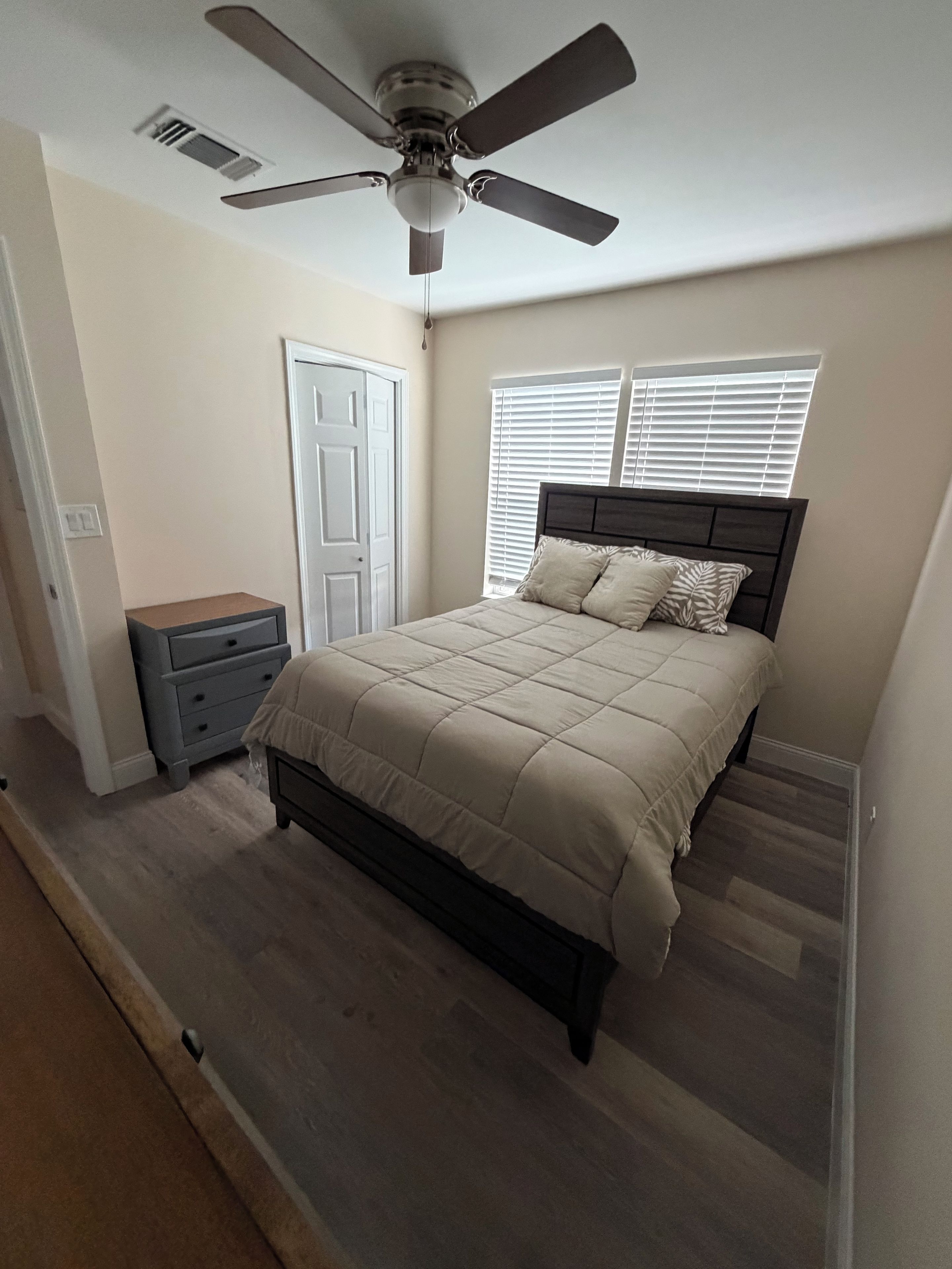 Un dormitorio con cama, mesita de noche, ventilador de techo y ventana con persianas.