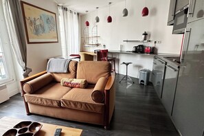 A modern living room with a brown sofa, a coffee table, and a kitchen area with a refrigerator, stove, and hanging pendant lights. - Cozy  Flat - La Fayette / Grands Boulevards (Paris)