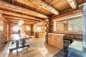A log cabin interior with a dining area, kitchen, and a large window overlooking the forest.