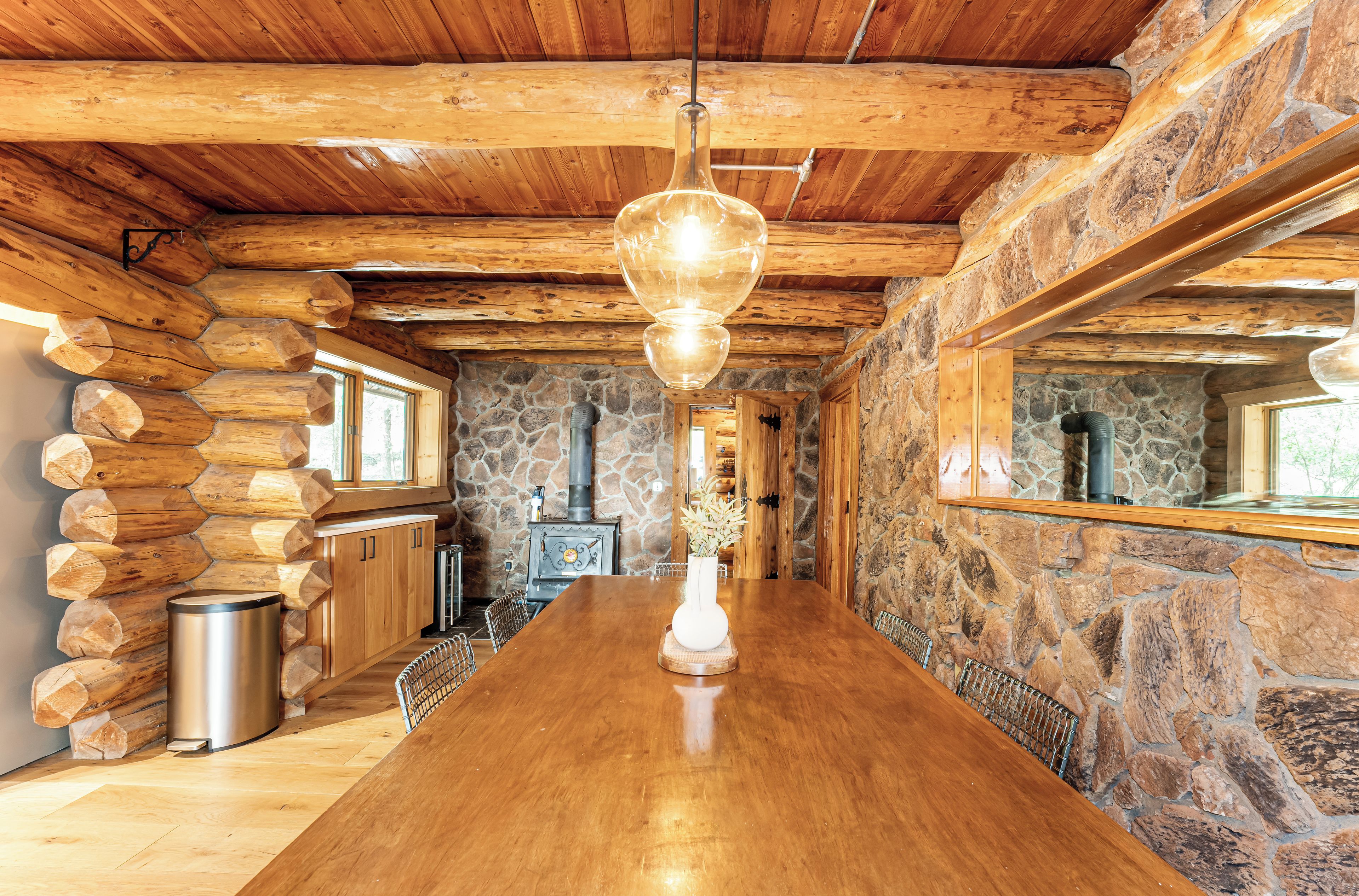 A log cabin interior with a wooden dining table, stone walls, and a hanging light fixture.