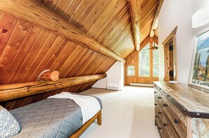 A room with a sloped ceiling, wooden walls, a bed, a dresser, and a painting.