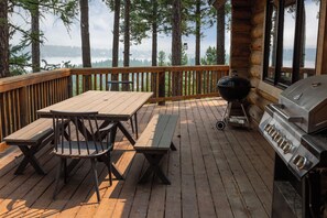 A wooden deck with picnic tables and a grill, overlooking a forested area and a body of water.