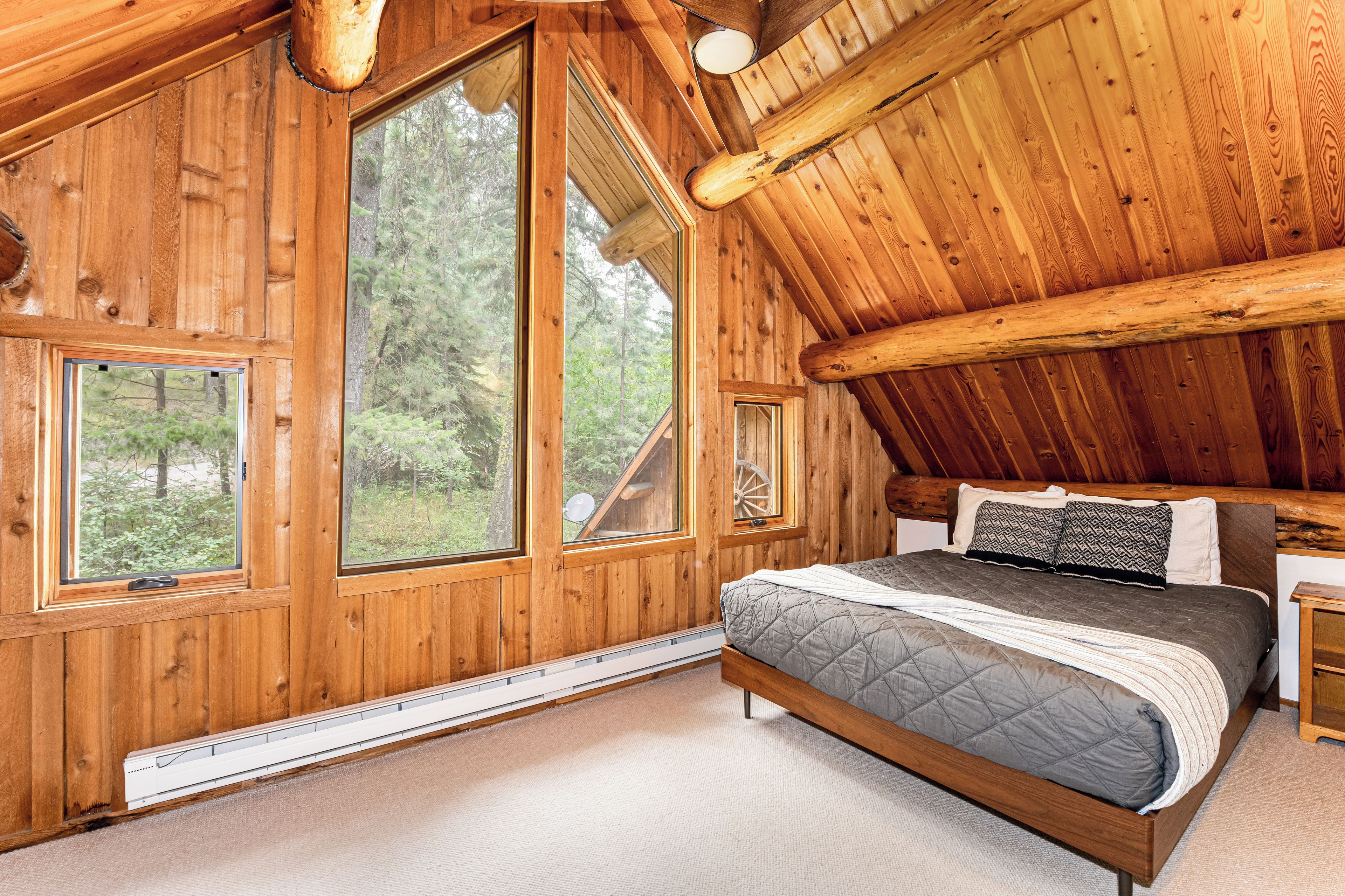 Quarto de cabana de madeira com uma cama grande, duas janelas e vista para a floresta.